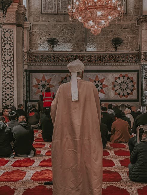 A group of people engaged in prayer within an ornately decorated mosque interior featuring beautiful patterns and a grand chandelier.